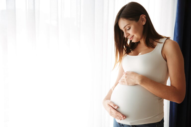 A pregnant woman stands by a window, gently cradling her belly and gazing outside with a serene expression.