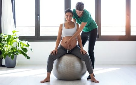 a woman standing behind another woman who is pregnant and sitting on a birth ball